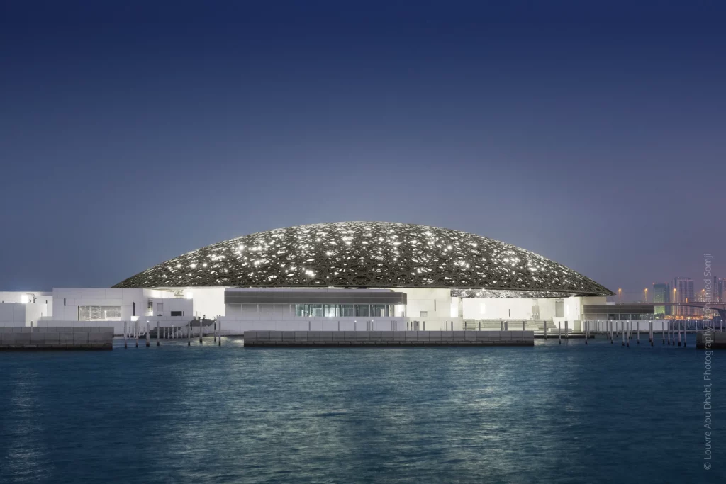 Vue extérieure nocturne du Louvre Abu Dhabi, avec la skyline d’Abu Dhabi en arrière-plan et la coupole illuminée reflétant sa lumière sur les eaux environnantes. Projet lumière conçu par 8’18’’, avec la participation de David Roger pendant 7 ans, des études à la phase chantier.